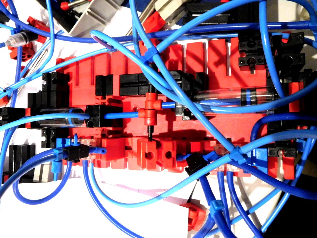 Pneumatikschaltung mit Umschalt-Triggerventilen (unten). Aus dem offnenen Schlauchstück (links unten) kommt gedrosselte Luft. Wird die Öffnung abgedeckt, baut sich Druck in einem Betätiger (blaues Element links auf der Bauplatte) auf, der ein Fischertechnik-Pneumatikventil (transparentes Element auf der Bauplatte rechts neben dem Betätiger) schaltet.